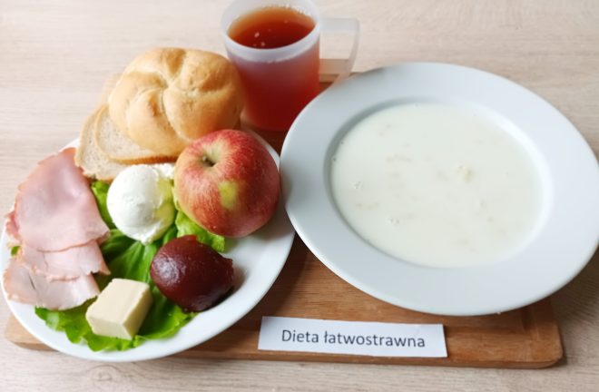 Na zdjęciu przedstawiono śniadanie serwowane w ramach diety łatwostrawnej, ułożone na jasnym, drewnianym blacie. Po lewej stronie znajduje się biały talerz z różnorodnymi składnikami: świeża bułka kajzerka i dwie kromki białego pieczywa, całe czerwono-zielone jabłko, dwa plasterki gotowanej szynki, plaster masła, kulka twarogu, gotowany burak oraz liść sałaty służący jako dekoracja. Po prawej stronie talerza umieszczono głęboki biały talerz z mlekiem i pływającymi w nim drobnymi płatkami – zapewne kaszą manną. W tle stoi przezroczysty plastikowy kubek z ciemną herbatą bez cytryny. Pod zestawem widoczna jest biała etykieta z czarnym napisem „Dieta łatwostrawna.