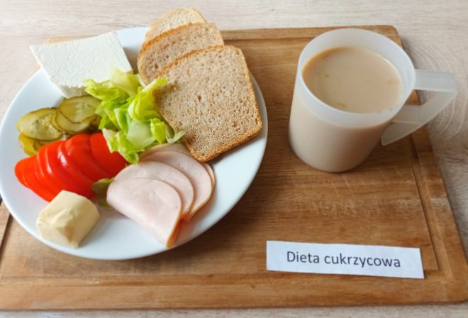 Śniadanie na drewnianej tacy. Na talerzu widoczne są trzy kromki pieczywa pełnoziarnistego, plasterki wędliny drobiowej, liście sałaty, plasterki pomidora i ogórka konserwowego, porcja białego sera i masła. Obok stoi kubek z kawą zbożową lub mlekiem. W dolnym prawym rogu etykieta „Dieta cukrzycowa”. Posiłek zbilansowany, lekkostrawny i odpowiedni dla osób z cukrzycą