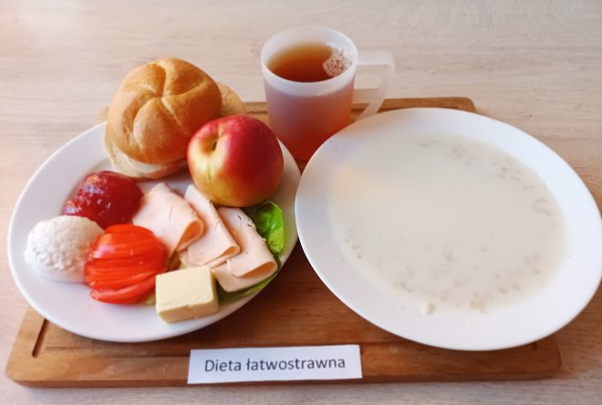 Na drewnianej tacy ustawiono posiłek śniadaniowy przeznaczony dla osoby na diecie łatwostrawnej. Na białym talerzu znajduje się okrągła bułka pszenna, jabłko, kilka plasterków delikatnej wędliny z kurczaka lub indyka, liść sałaty, plasterek sera żółtego, dwie cienkie plastry pomidora, porcja białego twarożku i łyżka galaretki owocowej. Obok talerza znajduje się biały głęboki talerz z mleczną zupą na bazie płatków, a także przezroczysty kubek z czerwoną herbatą lub naparem. Całość opisana etykietą „Dieta łatwostrawna”