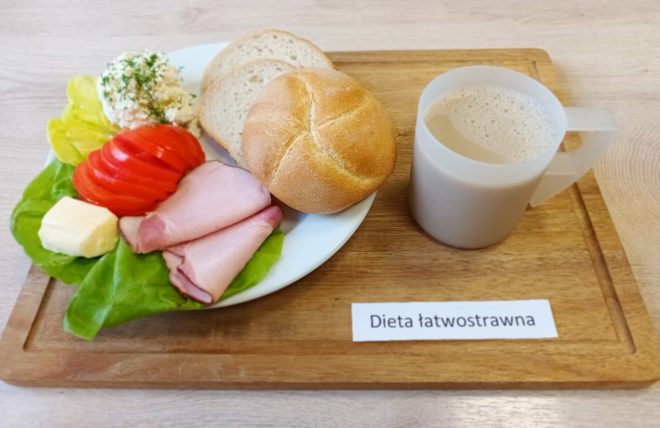 Śniadanie na jasnej drewnianej tacy. Na białym talerzu: liść sałaty, na nim plasterki szynki, masło, plasterki pomidora, sałata lodowa, porcja białego twarożku z koperkiem oraz dwa rodzaje pieczywa – pszenna bułka i kromki chleba mieszanego. Po prawej stronie plastikowy kubek z mlekiem lub napojem mlecznym. Pod posiłkiem kartka z napisem: „Dieta łatwostrawna”