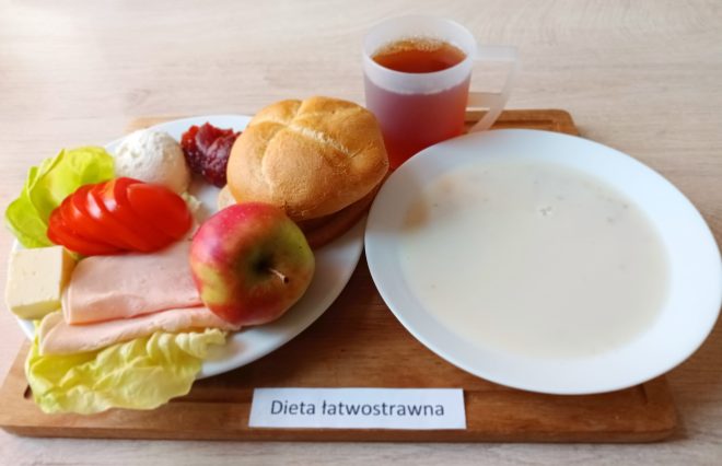 Na drewnianej tacy z podpisem "Dieta łatwostrawna" zaprezentowano zestaw śniadaniowy. Główne składniki znajdują się na dużym talerzu po lewej stronie i obejmują świeżą kajzerkę, plasterki pomidora, sałatę masłową, twarożek, plasterki chudej wędliny drobiowej, biały ser, cienki plasterek żółtego sera, porcję konfitury oraz jabłko z czerwono-zieloną skórką. Obok, na osobnym talerzu, znajduje się talerz z jasną zupą mleczną lub kleikiem. Zestaw uzupełnia kubek z herbatą o bursztynowym kolorze. Całość prezentuje się lekko, naturalnie i zgodnie z zasadami diety lekkostrawnej.