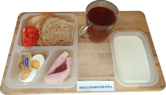 Na drewnianej tacy ułożono zestaw śniadaniowy w diecie podstawowej. W plastikowej tacko-tacy podzielonej na przegródki znajdują się: dwie kromki chleba razowego, plaster pomidora, jajko ugotowane na twardo przekrojone na pół, opakowanie masła oraz dwa zwinięte plastry szynki. W osobnych naczyniach: szklanka z ciemną herbatą oraz pojemnik z białym jogurtem naturalnym lub mlekiem. Pod tacką widoczny jest pasek z podpisem „DIETA PODSTAWOWA”