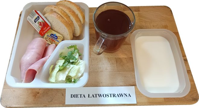 Zestaw śniadaniowy dla osób na diecie łatwostrawnej, ułożony na drewnianej tacy. W pojemniku podzielonym na trzy części umieszczono: dwie zwinięte plastry chudej szynki, porcję zielonej sałaty oraz trzy kromki białego pieczywa. W środku znajdują się również porcje dodatków w opakowaniach jednorazowych – masło oraz dżem truskawkowy. Po prawej stronie tacy ustawiono szklankę z ciemnym naparem herbaty oraz oddzielny pojemnik z białym płynem, prawdopodobnie mlekiem. Na tacy widnieje etykieta z napisem „DIETA ŁATWOSTRAWNA”