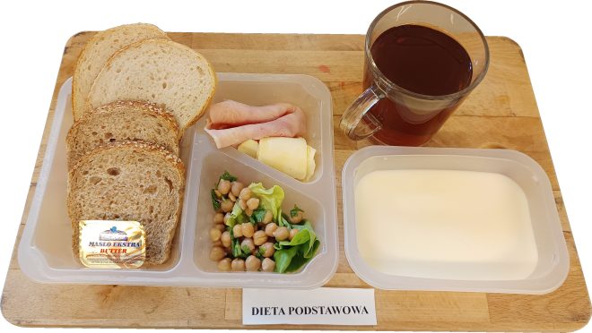 Na plastikowej tacy leży śniadanie w ramach diety podstawowej. W lewej przegródce znajdują się cztery kromki pieczywa – dwie z chleba jasnego i dwie z chleba wieloziarnistego. Obok leży porcja masła w zamkniętym opakowaniu. W górnej przegródce znajdują się dwa ruloniki wędliny: jeden z szynki i jeden z żółtego sera. Dolna przegródka zawiera sałatkę z ciecierzycy, zielonej sałaty i natki pietruszki. Do śniadania podano herbatę w przezroczystym kubku oraz porcję mlecznego deseru w prostokątnym pojemniku. Wszystko ułożone na drewnianym blacie z widoczną etykietą „Dieta podstawowa”