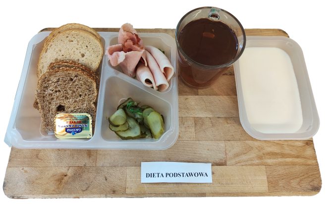 Śniadanie w diecie podstawowej podano w plastikowej, przegródkowej tacce, umieszczonej na drewnianej podstawce. W jednej przegródce znajdują się cztery kromki pieczywa – dwie jasne i dwie ciemne, razowe. W drugiej – plasterki chudej wędliny (szynka i indyk), a w trzeciej – plasterki ogórków konserwowych. Na pieczywie ułożono porcję masła w zamkniętym opakowaniu marki Łaciate. Obok tacki ustawiono szklankę z herbatą oraz dużą miseczkę z białym jogurtem naturalnym lub mlekiem. Całość opisana jako „Dieta podstawowa”