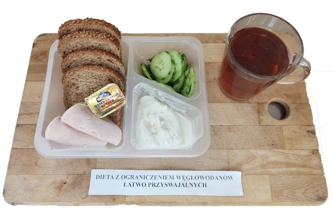 Na plastikowej tacce podanej na drewnianej desce umieszczono zestaw śniadaniowy w ramach diety z ograniczeniem węglowodanów łatwo przyswajalnych. Zestaw składa się z trzech kromek chleba razowego z ziarnami, dwóch plastrów chudej wędliny drobiowej, małego opakowania masła ekstra „Mlekovita”, porcji białego serka twarogowego oraz plasterków świeżego ogórka. Obok tacki stoi szklanka z przezroczystego szkła, wypełniona ciemnym naparem – prawdopodobnie herbatą bez cukru. Na desce widoczny jest wydrukowany pasek z opisem diety: „DIETA Z OGRANICZENIEM WĘGLOWODANÓW ŁATWO PRZYSWAJALNYCH”