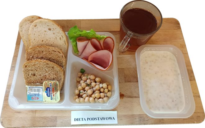 Na zdjęciu śniadanie w ramach diety podstawowej, serwowane na jasnej drewnianej tacy. W przezroczystym pojemniku po lewej stronie ułożono cztery kromki pieczywa: dwie z chleba razowego i dwie z jasnego pszennego. Obok znajdują się dwa opakowania tłuszczów: masło i miks tłuszczowy. Środkowa przegroda zawiera plasterki szynki z liściem sałaty oraz porcję gotowanej ciecierzycy. Po prawej stronie – przezroczysty kubek z herbatą plastikowy pojemnik z owsianką mleczną. Na środku tacki umieszczono karteczkę z napisem „DIETA PODSTAWOWA”.