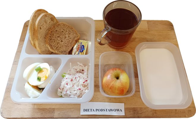 Na zdjęciu widoczny jest zestaw śniadaniowy podany na drewnianej tacy, oznaczonej etykietą „DIETA PODSTAWOWA”. W plastikowym pojemniku z czterema przegródkami umieszczono dwie kromki pieczywa pszennego i jedną kromkę pieczywa razowego, obok których znajduje się jednostkowe opakowanie masła marki Łaciate. W kolejnej przegródce znajdują się dwa plasterki jajka na twardo, ozdobione niewielką ilością drobno posiekanego szczypiorku. W trzeciej przegrodzie podano surówkę z białej rzodkwi i czerwonej rzodkiewki z dodatkiem zielonej cebulki – warzywa są starte na grubych oczkach, a całość wygląda świeżo i lekko wilgotno. Na tacy, poza głównym pojemnikiem, znajduje się także przezroczysty kubek z czerwonym napojem, najprawdopodobniej herbatą owocową. Obok ustawiono plastikowy pojemnik z całym jabłkiem w odcieniach czerwieni i żółci, a także prostokątne naczynie wypełnione białym płynem, który może być kefirem lub mlekiem
