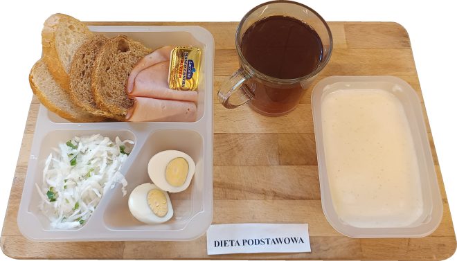 Śniadanie ułożone na plastikowej tacy z podziałem na przegródki, ustawionej na drewnianej desce. W górnej części znajdują się trzy kromki pieczywa: dwie jasne i jedna ciemna, plasterki chudej wędliny drobiowej oraz porcja masła w opakowaniu „Lactima”. W jednej z przegródek leży surówka z białej rzodkwi z dodatkiem szczypiorku, a obok dwa połówki gotowanego jajka na twardo. Po prawej stronie znajduje się kubek z ciemną herbatą w przezroczystym szkle oraz osobna miseczka z jasnym kleikiem mlecznym. Poniżej zestawu widnieje podpis: „DIETA PODSTAWOWA”