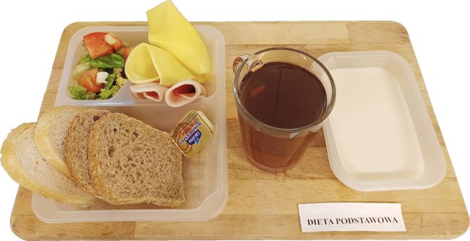 Zbilansowane śniadanie podane na drewnianej desce. W plastikowej tacce kilka kromek chleba pszennego i razowego, porcje żółtego sera i wędliny, małe opakowanie masła oraz miseczka z sałatką z pomidorów i sałaty. Obok tacki szklanka ciemnego napoju (prawdopodobnie herbata) oraz biała miseczka – prawdopodobnie zawierająca jogurt naturalny lub mleko. Poniżej etykieta: „DIETA PODSTAWOWA”