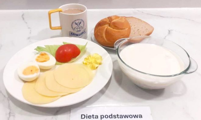 Śniadanie na jasnym blacie kuchennym. Na białym talerzu podano kilka plasterków sera żółtego, połówki jajka na twardo, liść sałaty, plaster pomidora oraz porcję masła. Obok znajduje się bułka pszenna typu kajzerka i kromka jasnego pieczywa. W przezroczystej miseczce ustawionej po prawej stronie widoczny jest biały napój mleczny, najprawdopodobniej kefir lub mleko. W tle ustawiona jest filiżanka z napojem – prawdopodobnie herbatą z mlekiem. Całość opisana jest karteczką z napisem „Dieta podstawowa”