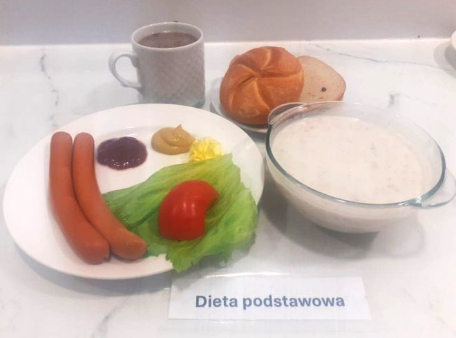 Zestaw śniadaniowy w ramach diety podstawowej na marmurowym blacie. Na talerzu ułożono dwa parówki, plaster pomidora i liść sałaty, a obok trzy sosy: ketchup, musztardę i porcję masła. W tle znajduje się kubek z kawą z mlekiem oraz dwa rodzaje pieczywa: bułka kajzerka i kromka chleba. Do zestawu dołączono także miskę z białą zupą mleczną z drobną kaszą. Całość oznaczona paskiem z napisem „Dieta podstawowa”