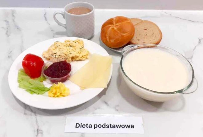 Na zdjęciu zaprezentowano śniadanie w ramach diety podstawowej, podane na marmurowym blacie. Na białym talerzu znajduje się porcja jajecznicy z koperkiem, dwa plasterki żółtego sera, listki sałaty, pół pomidora, porcja masła oraz mały pojemniczek z dżemem. Obok leżą dwa kawałki pieczywa – bułka i kromka chleba. Zestaw uzupełnia kubek z kawą z mlekiem oraz miska z białą zupą mleczną. Przed posiłkiem widnieje etykieta z napisem „Dieta podstawowa”