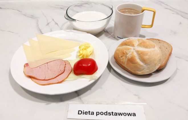 Śniadanie w ramach diety podstawowej, podane na marmurowym blacie. Na pierwszym talerzu znajdują się trzy trójkątne plasterki sera żółtego, dwa plasterki wędzonej szynki, liść sałaty, ćwiartka pomidora i porcja masła w formie dekoracyjnej rozety. Obok, na małym talerzyku, położono bułkę kajzerkę i kromkę chleba pszennego. W tle widoczna jest przezroczysta miseczka z mlekiem oraz biały kubek z kawą z mlekiem, z żółtym uchem. Przed posiłkiem znajduje się etykieta z napisem „Dieta podstawowa”