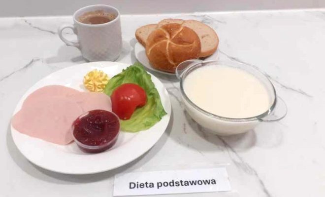 Drugie zdjęcie przedstawia zestaw śniadaniowy w diecie podstawowej. Na pierwszym planie talerz z dwoma plasterkami delikatnej szynki drobiowej, listkiem sałaty, ćwiartką pomidora, porcją masła w formie rozety oraz małą przezroczystą miseczką z konfiturą z czerwonych owoców. Obok znajduje się talerzyk z kajzerką i kromką chleba. W tle ustawiono miseczkę z białym mlekiem oraz filiżankę z kawą z mlekiem. Posiłek sfotografowano na jasnym marmurowym blacie. Przed jedzeniem umieszczona jest karteczka z napisem „Dieta podstawowa”