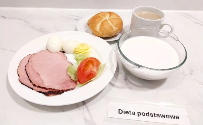 Śniadanie w ramach diety podstawowej podane zostało na białym talerzu ustawionym na marmurowym blacie. Zawiera kilka plasterków pieczonej szynki o różowej barwie i ciemniejszych brzegach, dwa obrane na twardo jajka kurze, dekoracyjnie uformowaną porcję masła oraz plasterek pomidora z listkiem sałaty. W tle widoczna jest okrągła bułka kajzerka ułożona na osobnym talerzyku. Do posiłku dołączono dwie porcelanowe filiżanki – w jednej znajduje się kawa z mlekiem lub inny napój mleczny, a w drugiej jasny płyn, prawdopodobnie mleko lub jogurt naturalny. Na stole przed talerzem widnieje etykieta z napisem „Dieta podstawowa”