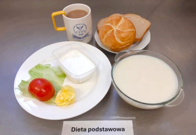 Szpitalne śniadanie w ramach diety podstawowej, sfotografowane na stalowym blacie. W skład zestawu wchodzą: sałata lodowa, plaster pomidora, masło w rozetce oraz pojemniczek z białym twarożkiem, podane na osobnym talerzu. Obok leży talerz z trzema kromkami pieczywa: jedna bułka typu kajzerka i dwie kromki chleba mieszanego. Do śniadania podano dużą miskę z mleczną zupą oraz kubek z napojem – najprawdopodobniej kawą zbożową z mlekiem. Kubek opatrzony jest logo uzdrowiska „Kryniczanka”