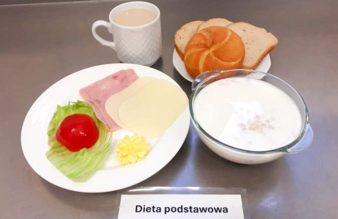 Na stalowym blacie widoczne śniadanie serwowane w ramach diety podstawowej. Na pierwszym planie znajduje się biały talerz z trzema plastrami – wędliną, żółtym serem oraz serem topionym, a obok porcja masła, plaster pomidora i liść sałaty lodowej. Z tyłu po prawej stronie stoi mała miseczka z dwoma kromkami chleba pszennego oraz jedną bułką kajzerką. Obok, w przezroczystej szklanej miseczce, znajduje się porcja płatków owsianych zalanych mlekiem. Całość dopełnia biały kubek z ciepłym napojem – prawdopodobnie kawa z mlekiem lub zbożowa kawa inka. Pod talerzami leży etykieta z napisem „Dieta podstawowa”
