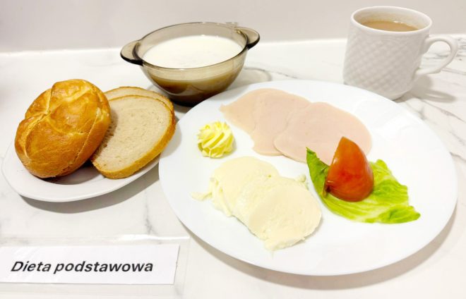 Na marmurowym blacie ułożono zestaw śniadaniowy w ramach diety podstawowej. Na dużym, białym talerzu znajdują się trzy plasterki delikatnej wędliny drobiowej, dwa plastry sera typu mozzarella, dekoracyjna rozetka masła oraz ćwiartka pomidora na liściu sałaty. Obok, na osobnym spodku, umieszczono świeżą bułkę kajzerkę i dwie kromki chleba pszennego. W tle stoi filiżanka z herbatą z mlekiem oraz szklana miseczka z białą zupą mleczną. Przed talerzem znajduje się etykieta z napisem „Dieta podstawowa”, wskazująca na rodzaj posiłku. Kompozycja na białym tle sprawia wrażenie czystości i staranności