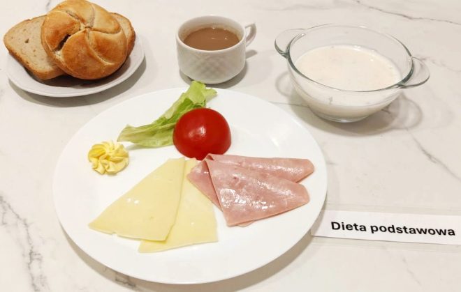 Na marmurowym blacie znajduje się klasyczny zestaw śniadaniowy w ramach „Diety podstawowej”. Główny talerz zawiera: dwa trójkątne plasterki żółtego sera, dwa plasterki szynki, połówkę czerwonego pomidora oraz liść sałaty. Obok – porcja masła dekoracyjnie uformowana. Na osobnym talerzyku: jasna kajzerka oraz kromki chleba mieszanego. Po prawej stronie filiżanka z kawą z mlekiem lub zbożowym napojem oraz miseczka z białą zupą mleczną lub kleikiem. Kompozycja prezentuje się schludnie i neutralnie, odpowiednia dla standardowej diety szpitalnej