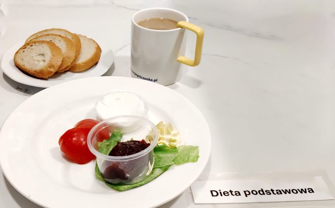 Na białym talerzu ułożono trzy grube plastry białej bułki, a obok na większym talerzu – pomidora w połówkach plasterków, liść sałaty, porcję dżemu w plastikowym kubeczku, porcję białego twarożku oraz rozetkę masła. Obok znajduje się kubek z kawą z mlekiem, z żółtym uchem. Na stole umieszczono etykietę z napisem „Dieta podstawowa”