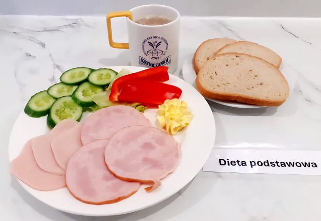 Śniadanie w diecie podstawowej, serwowane na białym talerzu. Kilka plasterków wędliny drobiowej, świeże warzywa (ogórek, papryka, sałata), porcja masła oraz pieczywo pszenne podane osobno na spodku. Obok filiżanka z napojem (prawdopodobnie kawa zbożowa lub herbata), kubek z logo ośrodka. Posiłek estetyczny, lekki i dostosowany do standardowej diety szpitalnej