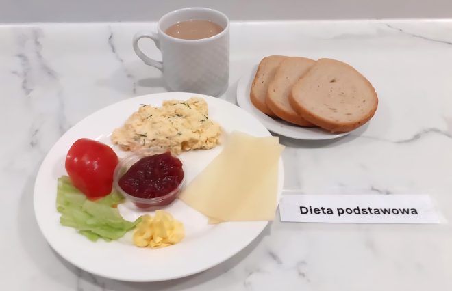 Na białym tle widać śniadanie wchodzące w skład „Diety podstawowej”. W centrum zdjęcia znajduje się talerz z porcją jajecznicy posypanej koperkiem, plastrem pomidora, liściem sałaty, dwiema cienkimi plastrowanymi żółtymi serami, porcją masła oraz niewielkim pojemniczkiem z konfiturą owocową. Po prawej stronie talerza leży talerzyk z trzema kromkami jasnego pieczywa pszennego. W tle widoczny jest biały ceramiczny kubek z kawą z mlekiem. Posiłek wygląda estetycznie i lekkostrawnie, zgodnie z założeniami diety podstawowej