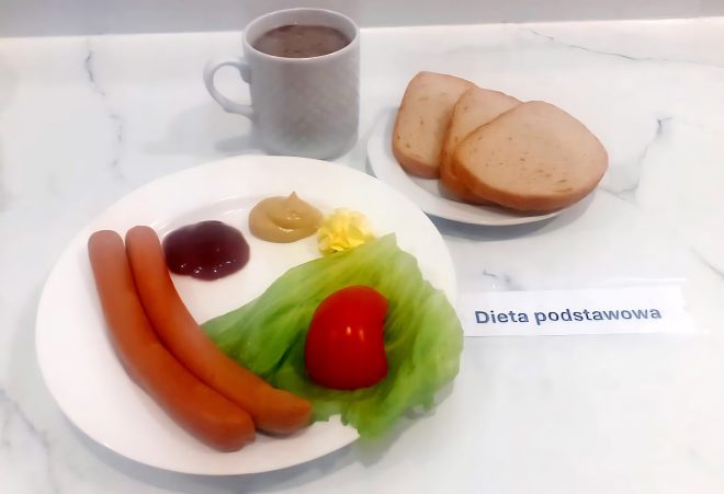 Śniadanie w ramach diety podstawowej, z etykietą „Dieta podstawowa”. Główny talerz zawiera dwa parówki, porcję ketchupu, musztardy i masła oraz dwa liście sałaty lodowej z połówką pomidora. W tle znajduje się osobny talerzyk z trzema kromkami jasnego pieczywa tostowego. Na środku, w białym kubku z uchem, podano napój – prawdopodobnie kawę zbożową lub herbatę z mlekiem. Kompozycja jest prosta i typowa dla podstawowego śniadania w placówkach leczniczych, zawiera źródła białka, warzywo, tłuszcze i węglowodany