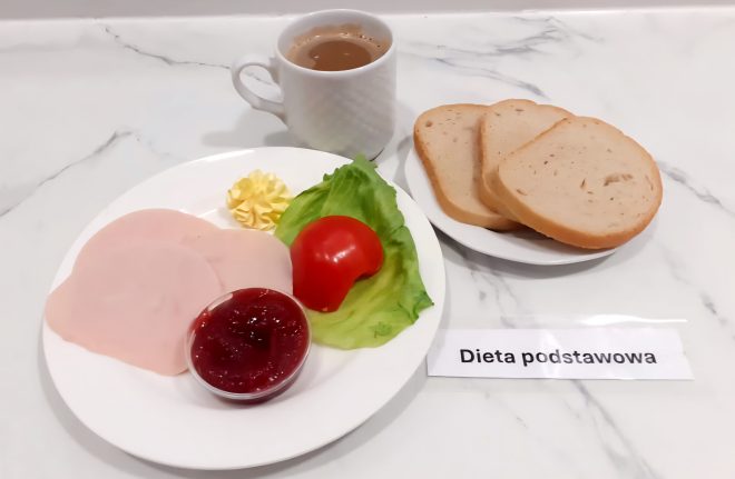 Śniadanie w ramach diety podstawowej, serwowane na marmurowym blacie. Na białym talerzu znajdują się trzy cienkie plasterki wędliny drobiowej, odrobina masła w formie rozety, kawałek sałaty lodowej, cząstka pomidora oraz mały pojemniczek z konfiturą truskawkową. Obok, na oddzielnym talerzyku, leżą trzy kromki jasnego pieczywa. W tle, po lewej stronie, widoczny jest biały ceramiczny kubek z kawą z mlekiem. Na blacie, przed talerzem, znajduje się etykieta z napisem „Dieta podstawowa”