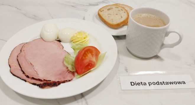 Na talerzu śniadaniowym, ustawionym na marmurowym blacie, znajduje się porcja zgodna z dietą podstawową. Zestaw zawiera dwa jajka ugotowane na twardo, trzy plasterki pieczonej szynki, dekoracyjnie uformowaną porcję masła oraz dodatek warzywny w postaci plastra pomidora z listkiem sałaty. W tle widoczny jest mniejszy talerzyk z dwiema kromkami jasnego chleba oraz biały porcelanowy kubek z napojem – prawdopodobnie kawa z mlekiem. Na pierwszym planie, przy dolnej krawędzi zdjęcia, znajduje się etykieta z napisem „Dieta podstawowa”, umieszczona w plastikowej koszulce. Ściana w tle pokryta jest jasnymi kafelkami, które nadają zdjęciu schludny i uporządkowany charakter