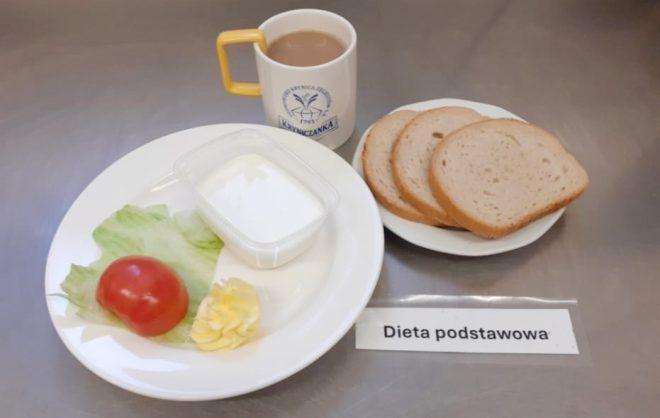 Na stalowym blacie stoi zestaw śniadaniowy oznaczony jako „Dieta podstawowa”. Na białym talerzu znajdują się: liść sałaty, połówka pomidora, porcja masła oraz przezroczyste pudełeczko z białym produktem nabiałowym (najprawdopodobniej jogurtem lub kefirem). Obok leży talerzyk z trzema kromkami białego pieczywa. Do śniadania podano gorący napój w kubku z logo „Kryniczanka” – najpewniej kawa z mlekiem lub zbożówka. Przed posiłkiem umieszczona jest etykieta „Dieta podstawowa”