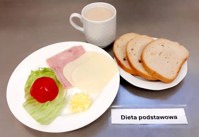 Drugie zdjęcie ukazuje zestaw śniadaniowy w ramach tej samej diety. Na głównym talerzu znajdują się dwa plastry wędliny (najprawdopodobniej szynki i indyka), dwa plasterki sera, liść sałaty lodowej z cząstką pomidora oraz porcja masła w formie dekoracyjnej rozetki. Obok, na oddzielnym talerzyku, ułożono trzy kromki białego pieczywa. Całość uzupełnia kubek z jasnym napojem, prawdopodobnie kawą z mlekiem lub zbożową, ustawiony w tle. Posiłek oznaczono etykietą „Dieta podstawowa”, umieszczoną na połyskującym, metalicznym blacie