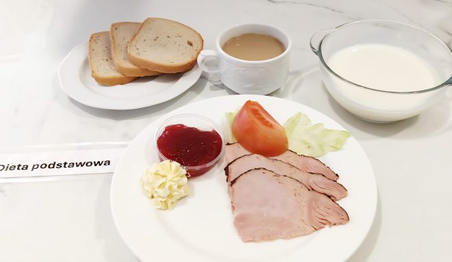 Zestaw śniadaniowy w ramach diety podstawowej, podany na marmurowym blacie. W centrum białego talerza znajdują się plasterki pieczonego schabu, ćwiartka pomidora, liść sałaty, odrobina masła oraz porcja czerwonej konfitury. Z lewej – talerzyk z trzema kromkami jasnego pieczywa tostowego. W głębi: biały porcelanowy kubek z kawą z mlekiem oraz szklana miska z mlekiem. Przed talerzem widnieje etykieta z napisem „Dieta podstawowa”