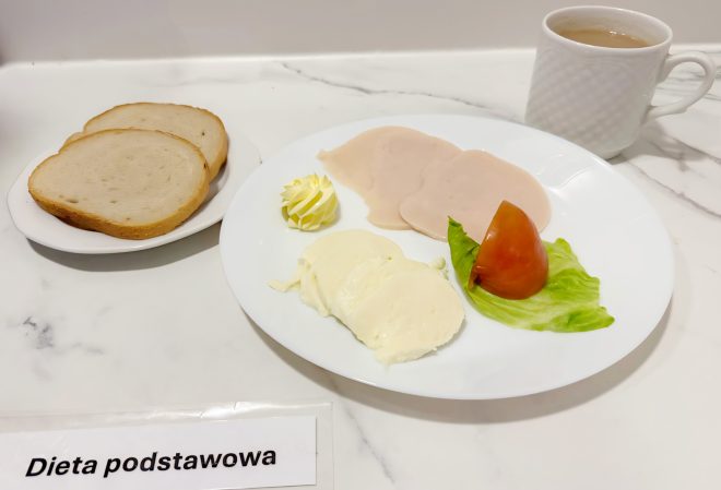 Na drugim zdjęciu przedstawiono śniadanie w ramach diety podstawowej, serwowane na białym blacie. Na dużym talerzu leżą dwa plasterki delikatnej wędliny drobiowej, dwa plastry sera typu mozzarella, porcja masła w dekoracyjnej formie rozetki oraz kawałek pomidora ułożony na listku sałaty. Obok na osobnym spodku znajdują się dwie kromki chleba pszennego. W tle po prawej stronie widoczna jest filiżanka z gorącą herbatą z mlekiem. Na dole zdjęcia leży etykieta z opisem „Dieta podstawowa”