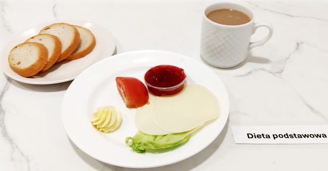 Śniadanie podane na marmurowym blacie z podpisem „Dieta podstawowa”. Na małym talerzyku znajdują się cztery kromki białego pieczywa. Na głównym talerzu obok ułożono dwie plastry sera żółtego, ćwiartkę pomidora, liść sałaty, rozetkę z masła oraz porcję dżemu truskawkowego w plastikowym kubeczku. Towarzyszy temu biały kubek z herbatą z mlekiem