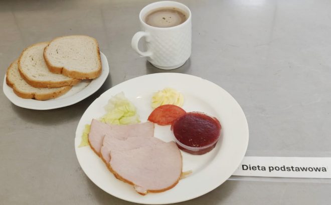 Na śniadaniowym talerzu ułożono trzy plastry pieczonego schabu, porcję sałaty lodowej, plaster pomidora oraz porcję masła i galaretki owocowej w małym, przezroczystym pojemniczku. Obok znajduje się talerzyk z trzema kromkami jasnego chleba. Z tyłu biały kubek z kawą z mlekiem. Całość ustawiona na stalowym blacie, z etykietą „Dieta podstawowa”