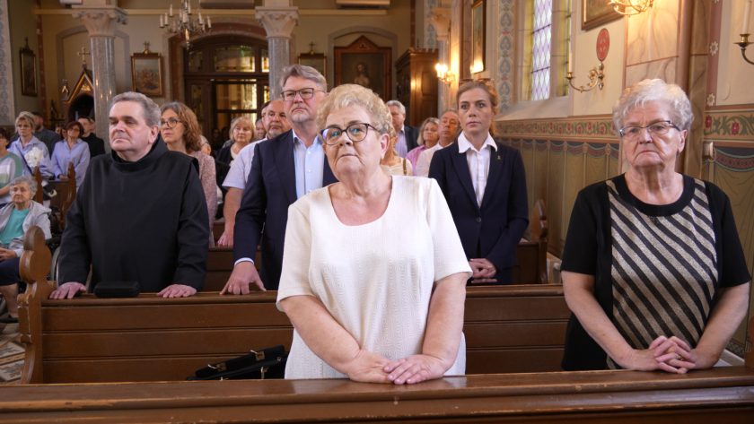 Wnętrze kościoła wypełnione wiernymi. W pierwszym rzędzie kilka osób – kobieta w białej sukience, duchowny bonifraterski, mężczyzna w garniturze i inne osoby – stoją w postawie modlitewnej. Tło tworzą bogato zdobione ściany i kolorowe witraże.