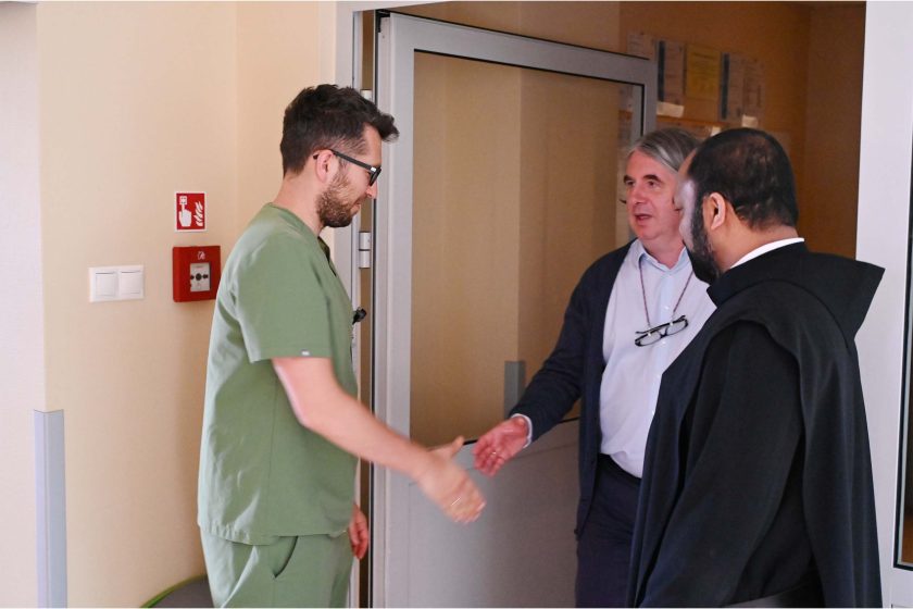 Mężczyzna w zielonym uniformie wita się z osobą w swetrze, obok której stoi brat zakonny. Spotkanie odbywa się przy wejściu do sali szpitalnej.