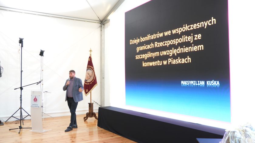 Prelegent Maksymilian Kuśka stoi z mikrofonem przed dużym ekranem prezentacyjnym z tytułem wykładu: „Dzieje bonifratrów we współczesnych granicach Rzeczpospolitej ze szczególnym uwzględnieniem konwentu w Piaskach”. Obok niego stoi sztandar Bonifraterskiego Centrum Medycznego, a całość odbywa się w jasnym namiocie konferencyjnym.