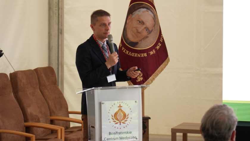 Prelegent przemawia przy mównicy z logo Bonifraterskiego Centrum Medycznego, w tle sztandar z wizerunkiem św. Benedykta Menni.