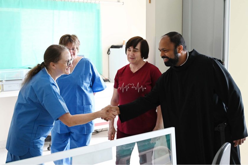 Brat zakonny w czarnym habicie uśmiecha się i podaje rękę jednej z trzech kobiet w stroju medycznym. Spotkanie odbywa się w jasnym pomieszczeniu szpitalnym.