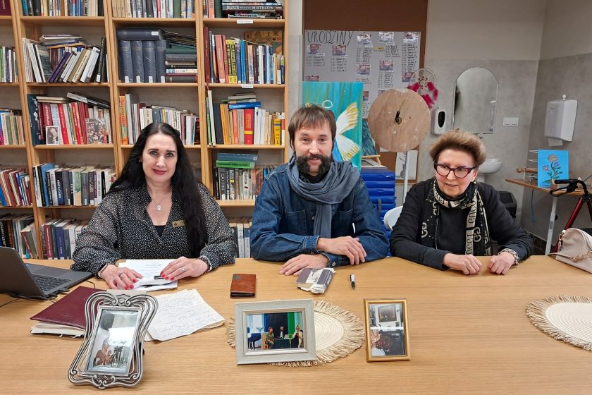 Portretowe zdjęcie prowadzących – troje dorosłych siedzi przy stole w sali bibliotecznej. Uśmiechnięci, patrzą w stronę aparatu. Za nimi regały pełne książek i dekoracje. Na stole kilka ramek ze zdjęciami oraz notatki, co sugeruje charakter spotkania literackiego lub wspomnieniowego