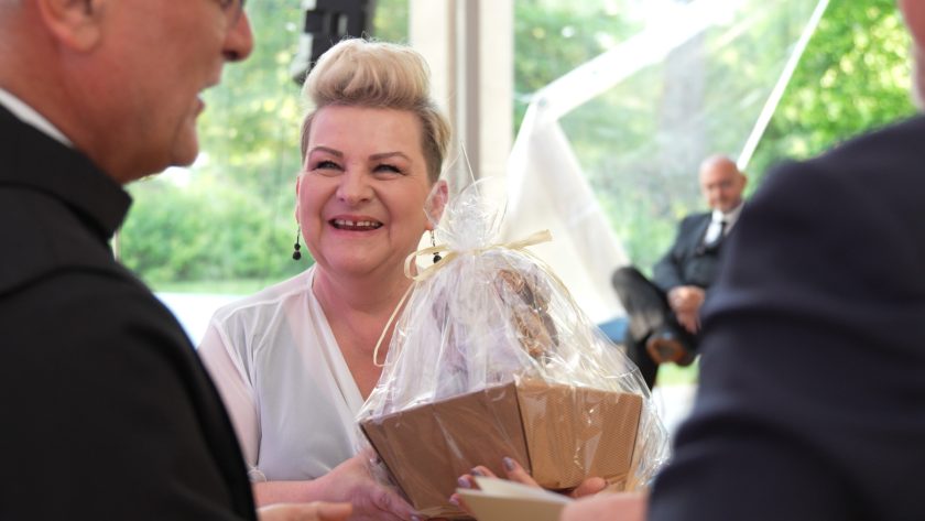 Uśmiechnięta kobieta o jasnych włosach i eleganckim uczesaniu odbiera prezent zapakowany w przezroczystą folię z rąk osoby spoza kadru. W tle widać rozmytą sylwetkę mężczyzny siedzącego na krześle oraz zielone drzewa za przezroczystą ścianą namiotu.