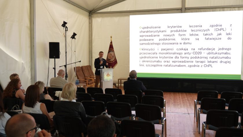 Sala konferencyjna z uczestnikami szkolenia; na ekranie prezentacja dotycząca kryteriów leczenia i refundacji leków.