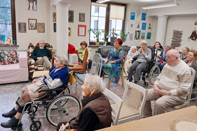 Wnętrze przytulnej świetlicy lub sali dziennego pobytu. Grupa starszych osób, część na wózkach inwalidzkich, siedzi w półkolu i skupia wzrok w jednym kierunku – prawdopodobnie w stronę prelegentów lub artystów. W tle jasne ściany ozdobione są dziecięcymi rysunkami i portretami, a z parapetów spoglądają doniczkowe kwiaty. Atmosfera skupienia i zainteresowania