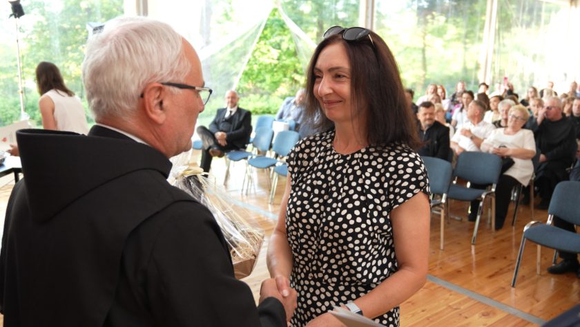 Zakonnika w habicie bonifraterskim wita uśmiechnięta kobieta w sukience w biało-czarne grochy. Uściski dłoni odbywają się na tle sali pełnej gości siedzących na krzesłach w jasnym, przeszklonym wnętrzu otoczonym zielenią.