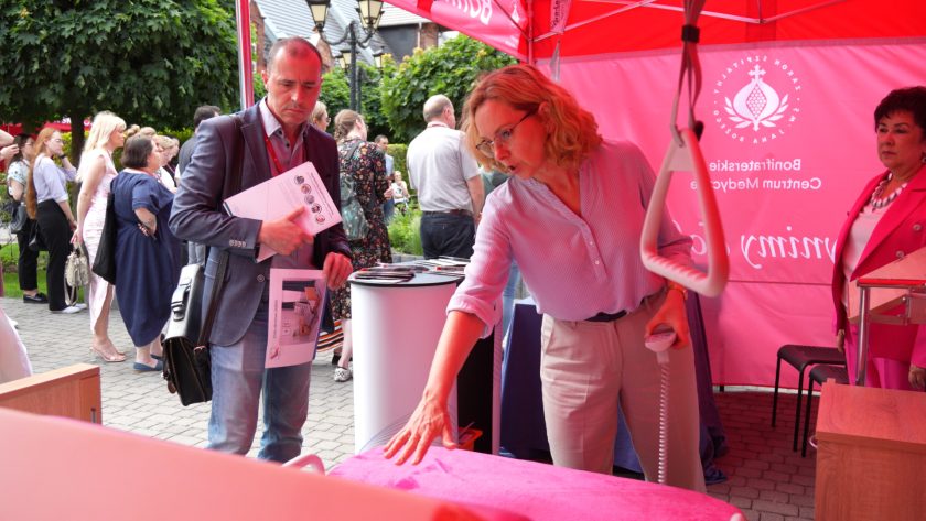 Kobieta prezentuje funkcje łóżka medycznego pod namiotem wystawienniczym, mężczyzna obok trzyma ulotki.