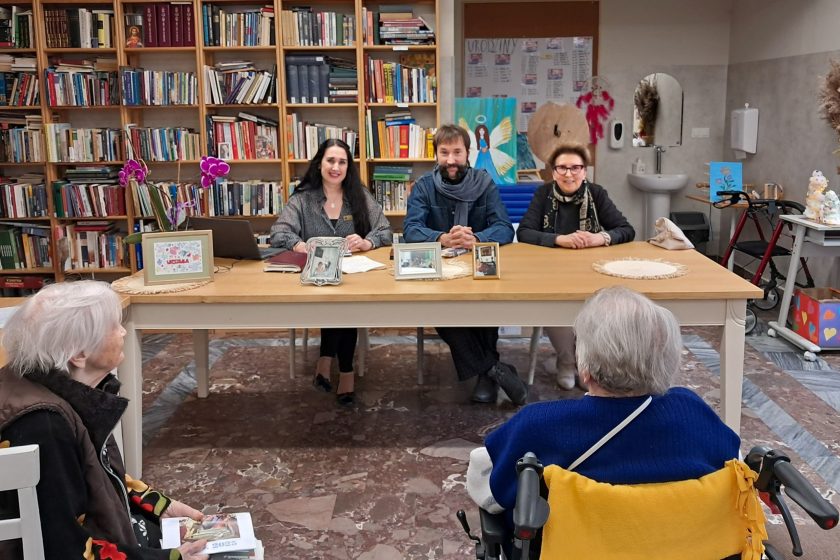Portretowe zdjęcie prowadzących – troje dorosłych siedzi przy stole w sali bibliotecznej. Uśmiechnięci, patrzą w stronę aparatu. Za nimi regały pełne książek i dekoracje. Na stole kilka ramek ze zdjęciami oraz notatki, co sugeruje charakter spotkania literackiego lub wspomnieniowego