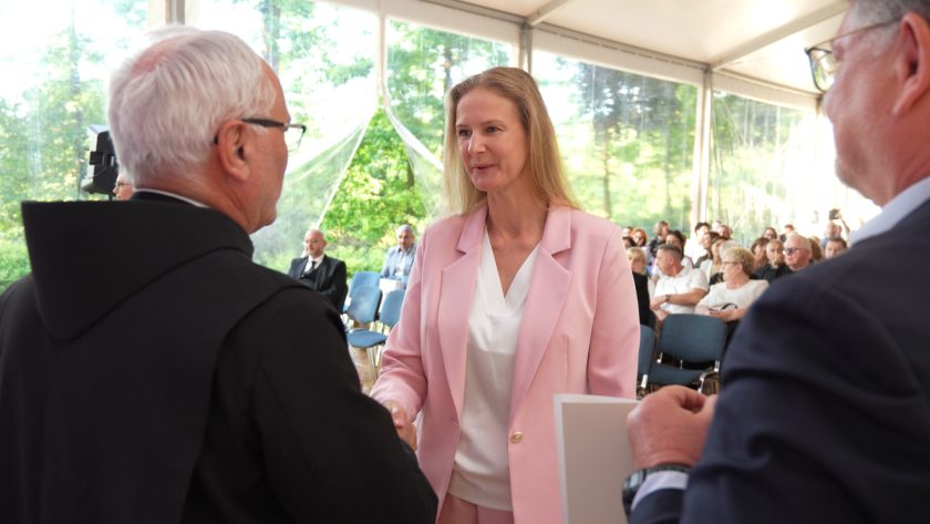 Młoda kobieta w różowym garniturze odbiera gratulacje i dyplom z rąk bonifratra. Uśmiecha się uprzejmie, trzymając dokument. W tle widać zgromadzonych gości i naturalne światło wpadające przez przezroczyste ściany namiotu.