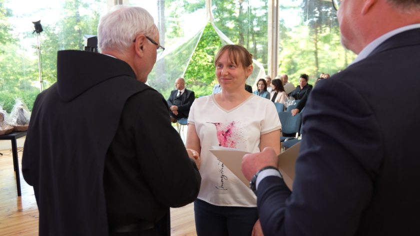 Kobieta w białej bluzce z kolorowym nadrukiem ptaka odbiera dyplom i uścisk dłoni od zakonnika. Uśmiecha się ciepło, a za nią, na tle sali, widać innych gości, przezroczyste ściany i drzewa na zewnątrz.